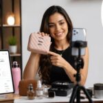 Mulher sorridente grava vídeo mostrando uma pequena bolsa rosa para o celular apoiado em um tripé com ring light.